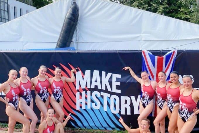 GB Artistic swimmers in costumes in front of a 'Make History' display poolside
