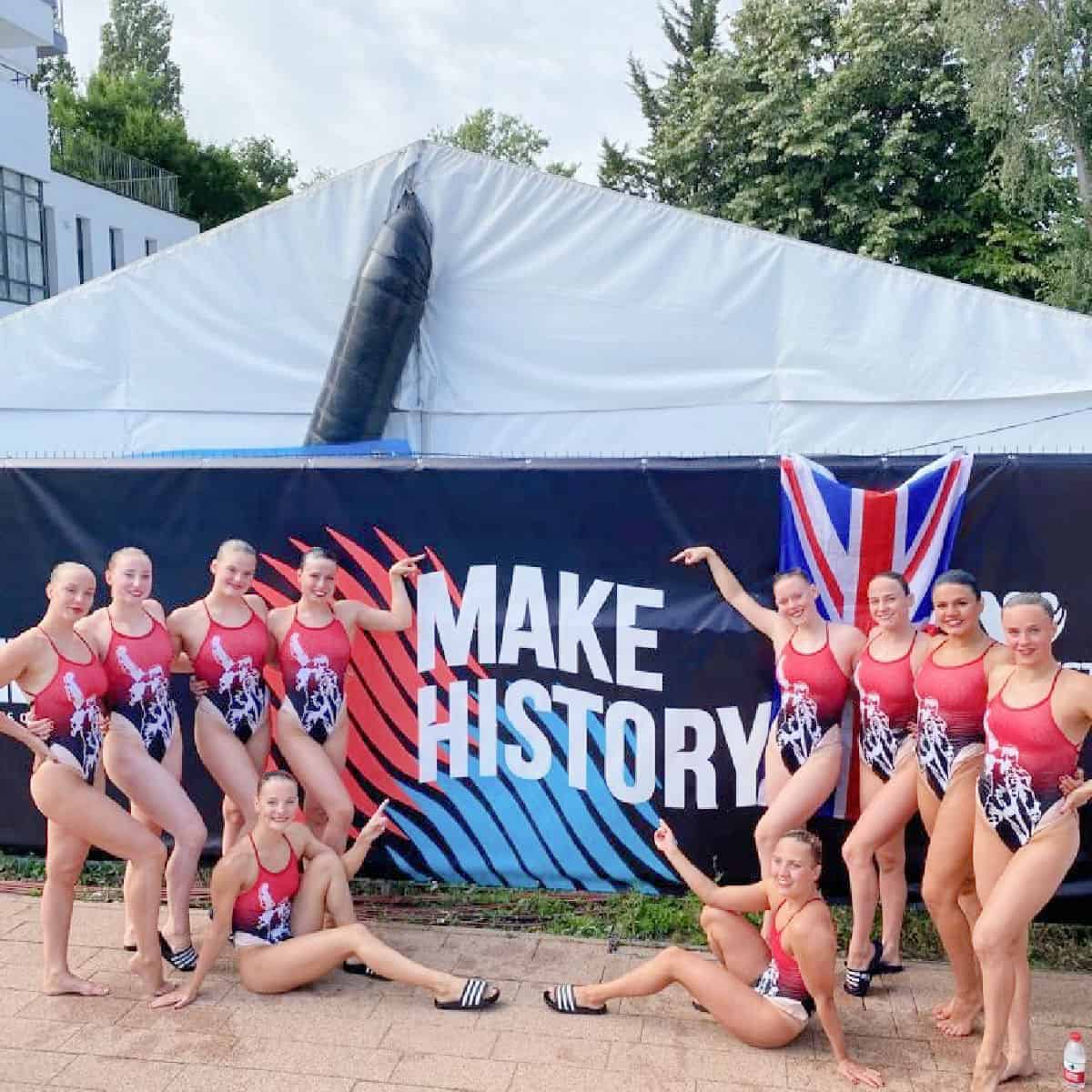 GB Artistic swimmers in costumes in front of a 'Make History' display poolside