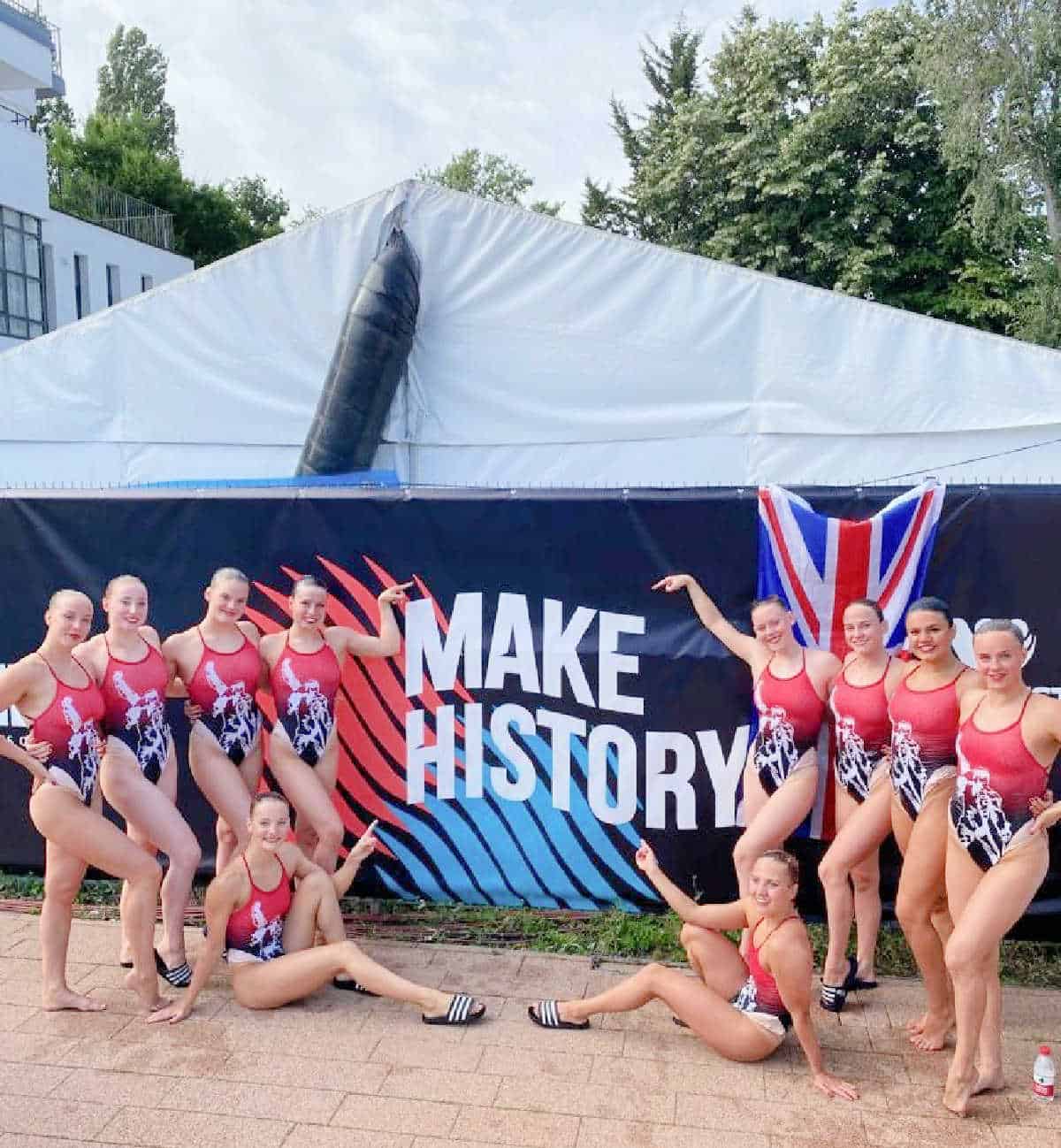 synchronised swimmers out of the water posing in front of a 'Make History' sign