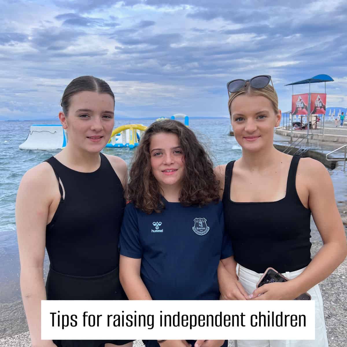 3 sisters against a backdrop of a black, one blond with sunglasses on her head, one brunette with hair tied back, the smallest in the centre with long brown curly hair wearing a navy everton shirt