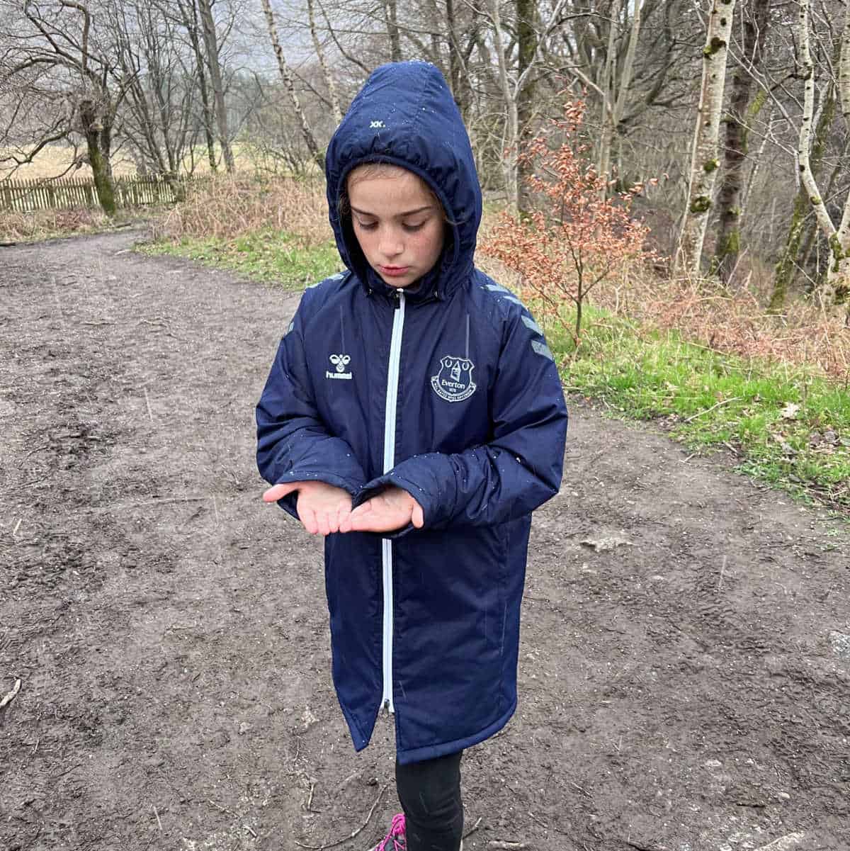 a child outside in the woods wearing a navy coat with her hood up, looking at her hands with a puzzled expression.