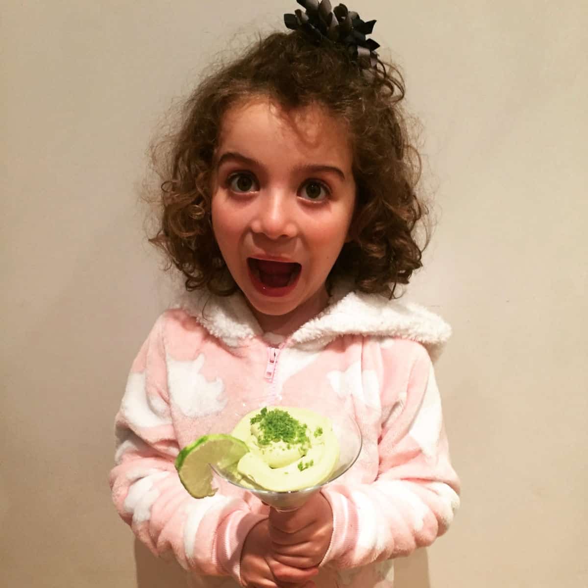 girl with curly brown hair and a pink and white fluffy top holding a green mousse in a glass with a slice of lemon pulling a funny face for the camera