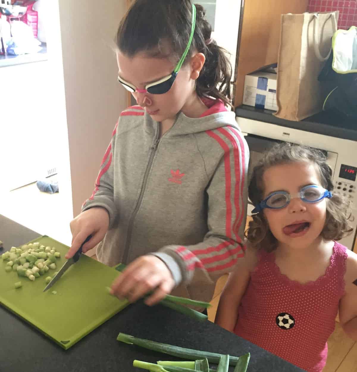 one young girl chopping spring onions on a green chopping board and one girl sticking her tongue to the side of her mouth. Both are wearing goggles in the kitchen.