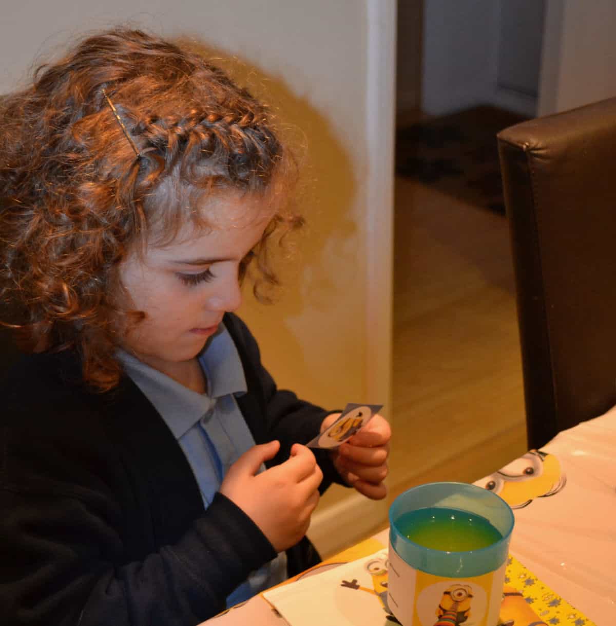 curly haired girl wearing a blue school uniform sticking minion stickers and drinking juice at a table