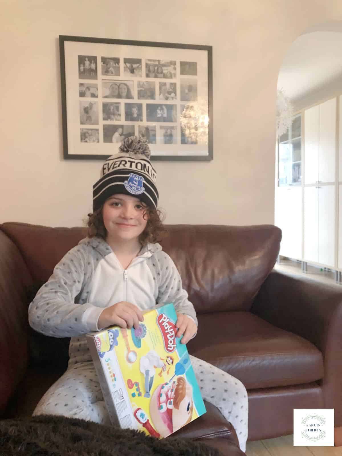 girl wearing a grey and white onesie and an everton bobble hat opening a box of playdoh