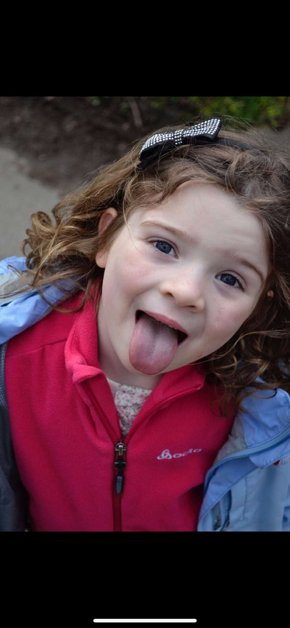 young girl with brown curly hair looking very happy and sticking her tongue out at the camera